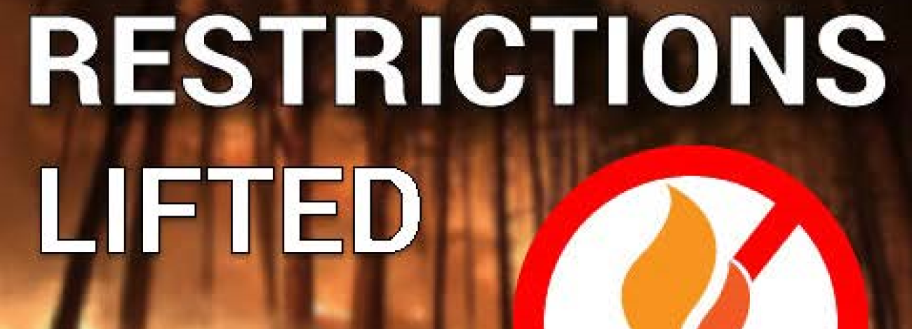 A wooded area with flames burning among the trees. Bold text across the image reads “Fire Restrictions Lifted.” In the bottom right corner, a graphic shows a red circle with a slash over a flame icon.