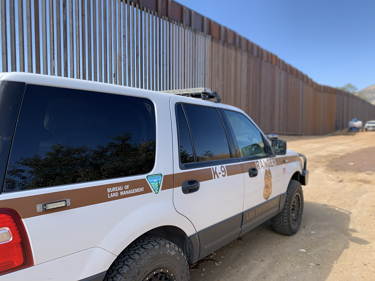 A BLM Law Enforcement vehicle (designated with the BLM logo and as a "K9 Vehicle) is parked by a tall, slatted wall on a dirt road.
