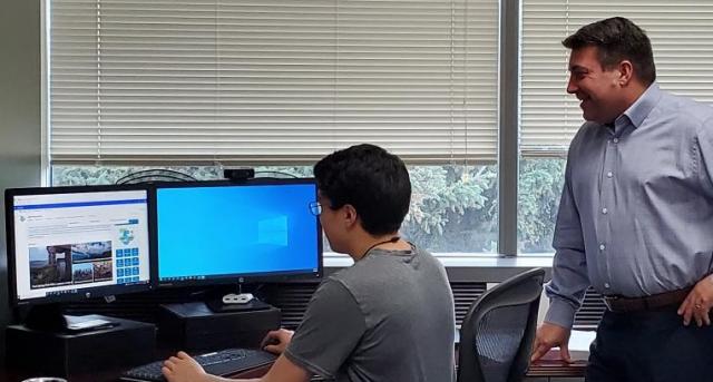 A man looks over the shoulder of another man working on his computer. 