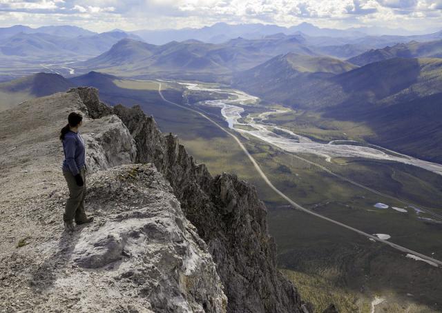 A hiker looks out from a craggy limestone peak over a braided river, highway, and pipeline