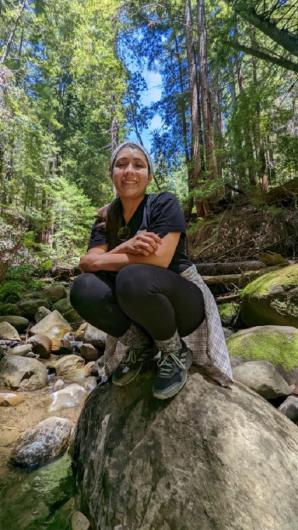 A woman crouches down on a boulder in a stream in a forest. 