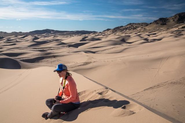 Photo shows woman in blue cap, sunglasses, orange shirt, and dark jeans, with a camera around her neck, sitting on a sand dune and surrounded by sand dunes and blue skies.
