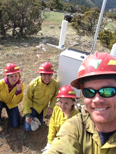 A person takes a selfie with three kids. All are wearing yellow firefighting shirts and red hardhats.