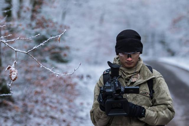 The photo shows a man facing the camera in a stocking cap, glasses, and winter fatigues as he stands near an ice-covered tree branch and looks down into the viewfinder of his own video camera.  
