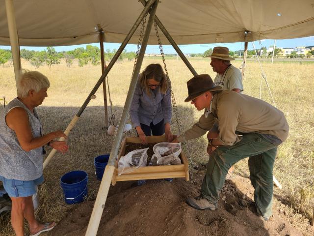 A photo of people conducting an archaeological dig outdoors with a pop up tent overhead.