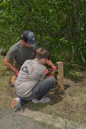 Two people use a drill to attach a campsite number to a wooden post.