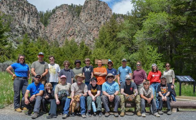 A group of about 20 kids and adults gather for a photo in front of scenic steep cliffs and green trees.