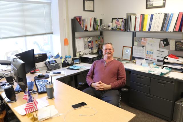 A man poses, smiling, at his desk.