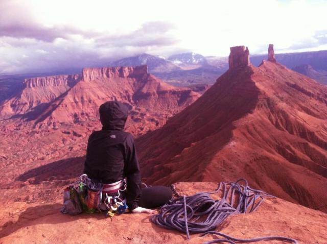 This photo shows a lone climber sitting next to a pile of climbing rope on the edge of a cliff looking out towards eroding red sandstone cliffs that stand out majestically against the cloudy sky. 