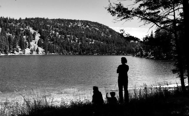 The black and white photo shows an adult standing next to two sitting children. They are on the shore of a small lake with a tree-covered hill in the background.