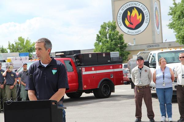 A Caucasian man in a blue polo shirt with the BLM logo on it gives a speech behind an outdoor podium.