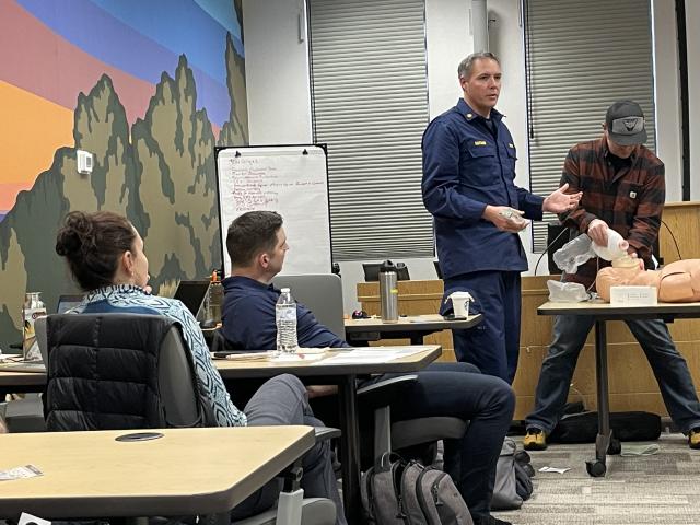 A white man in dark blue stands and speaks to a classroom while a person next to him pours water over a medical dummy.