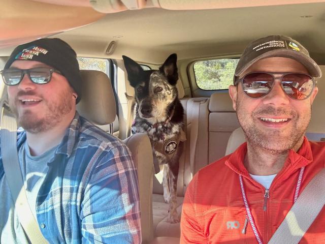 Photo shows two men sitting in a car with seat belts on, both are wearing hats and sunglasses; the man on the left is in a blue plaid shirt and the one on the right is in a bright orange pullover. A black and white dog is sitting in the backseat looking straight ahead.