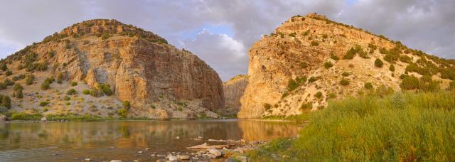 A serene landscape featuring two rocky cliffs surrounding a calm river. The cliffs are sunlit with patches of vegetation, while green reeds line the water's edge. Partly cloudy skies complete the natural scene.