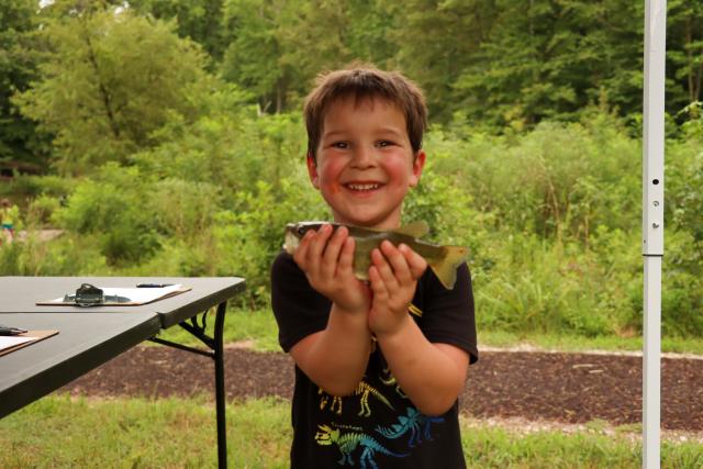 A young boy smiles widely as he holds a small fish with both hands in front of him, displaying it for the camera at the most recent FishingCommunity.org event at Meadowood SRMA.