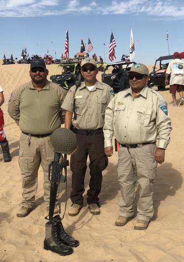 Three men stand solemnly behind military style boots, helmet and rifle..