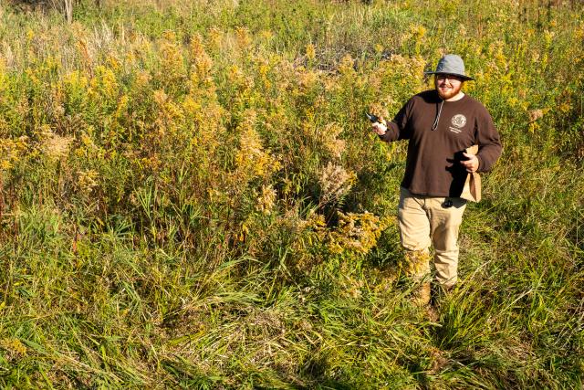 Person wearing a brown shirt and hat stands in a field of grass and tall plants.