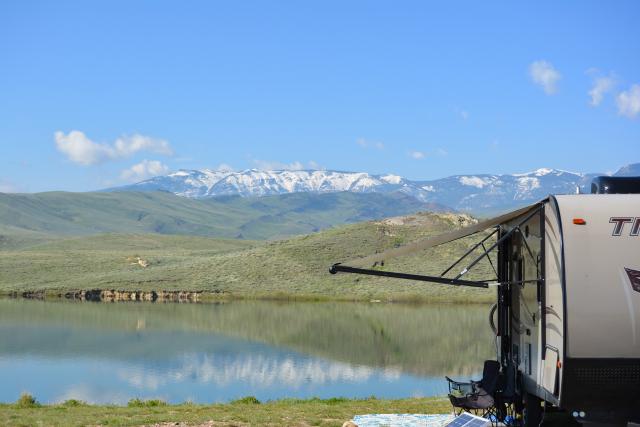 A camper is parked in a campground on the shore of a lake. Snow-topped mountains in the distance are reflected in the lakewater.