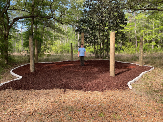 A teenaged boy stands in between four large posts in a hammock area he created with wooded area seen behind him. 