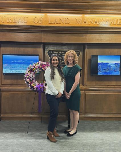 Vanessa Lacayo and Tracy Stone-Manning stand facing the camera in front of a flower wreath adorning the offices of the Bureau of Land Management in the Stewart Lee Udall Building in Washington, D.C.