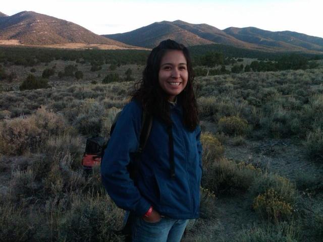 Vanessa Lacayo, smiling and wearing a blue jacket, stands in a field of sagebrush. Behind her is a line of small mountains, brownish green in color.