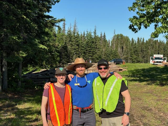 Three Caucasian adults in hats and work vests post smiling next to conifers and piled logs