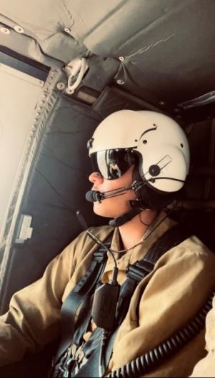 A Caucasian person in a white helmet with a black visor and mic attached and a brown flight suit sits inside an aircraft looking out the window.