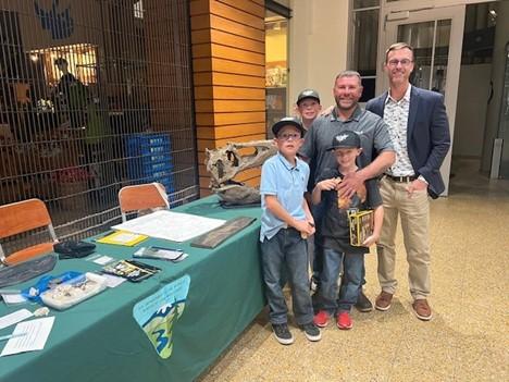 Two men and three boys in baseball caps pose in front of a table holding a model T. rex skull. 