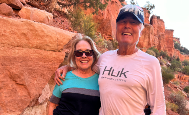 A couple stand on a trail in front of red rocks. 