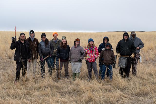 A row of people wearing cold weather gear stand in a grassy area smiling at the camera. 