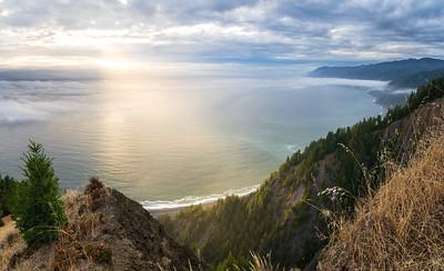 A sunset over  the Pacific Ocean with a steep forested ridge winding from foreground to back ground