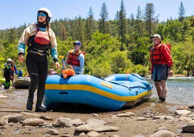 Three rafters in white water gear exit on a river beach in a forest.