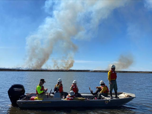 BLM firefighters are seen on a boat in the foreground, with smoke rising above the land across the water in the distance