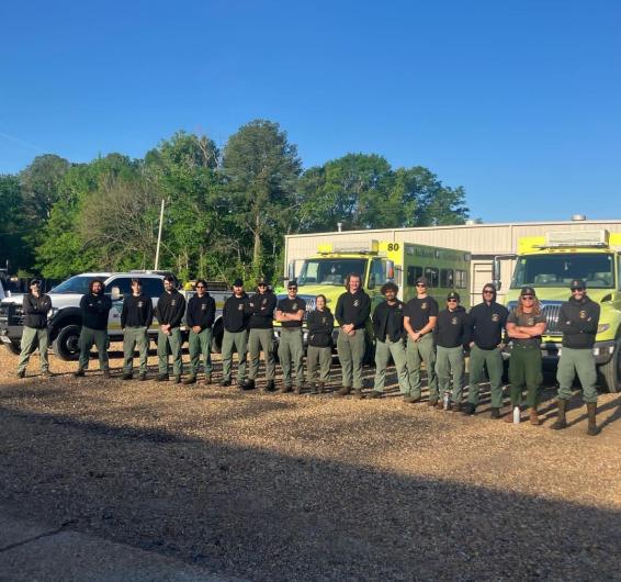 The Jackson Hotshots team is seen lined up posing for the camera, with a building and vehicles behind them. 