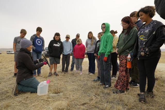 A group of young people watch and listen as a BLM employee shows them how to plant a sagebrush seedling.