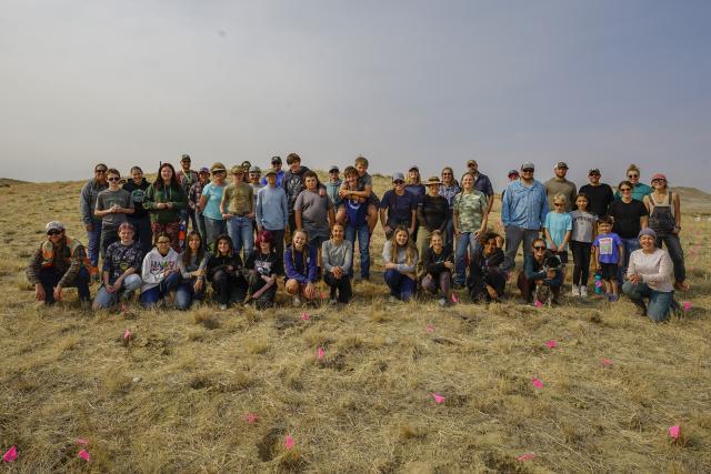 A large group of students and adult pose for a photo in an open, grassy landscape.