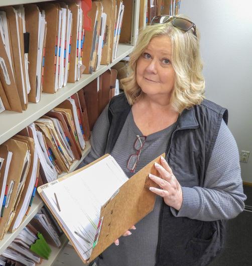 A woman stands next to a file cabinet packed with file folders, holding a file folder open.