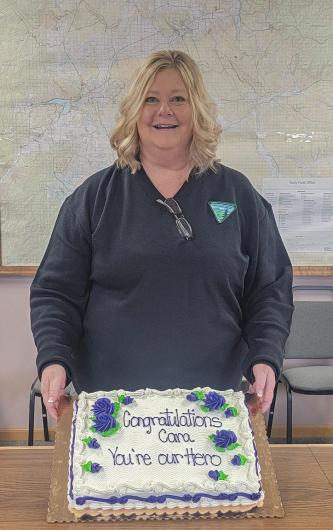 A woman poses for a picture behind a large cake that reads "Congratulations Cara You're Our Hero" with frosting.