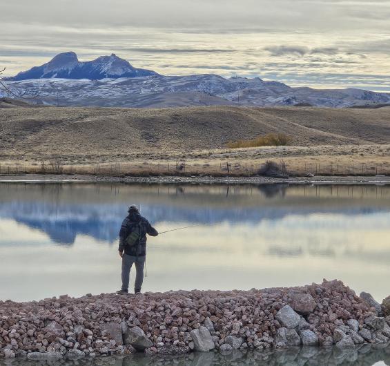 A person fishes from a rock jetty surrounded by the water of a scenic reservoir with partially snow-covered mountains for the backdrop.