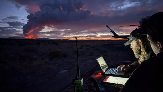 Two caucasian men's faces illuminated by the glow of a laptop screen with a glowing pink sunset and fire on the horizon