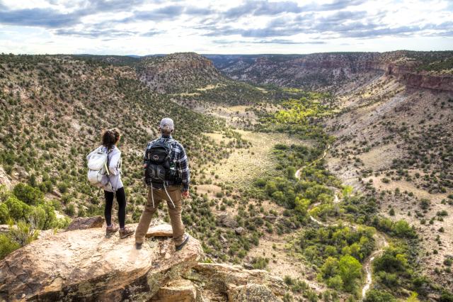 Two people with backpacks stand on rocks overlooking a portion of the Dominguez-Escalante National Conservation Area.