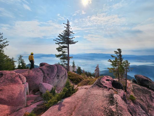 A person in a yellow shirt and hardhat walks over granite boulders towards a mountain vista