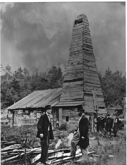 A photo from the 1860s showing two men standing in front of a wooden structure that contains the first oil well drilled in America. 