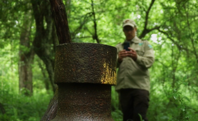 A metal well pipe sticks out of the ground in the foreground, as we see a man taking a picture of it, out of focus, in the background. 