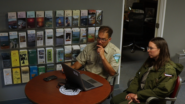 A man and woman, both in uniforms, sit at a table looking at a laptop screen. 