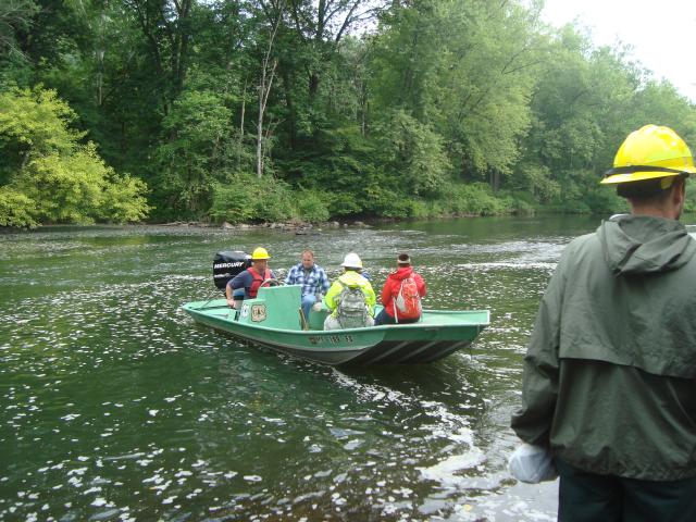 Three men on a motorboat approach the shore of an island in a river, as we see the back of a man standing on the shore awaiting them.