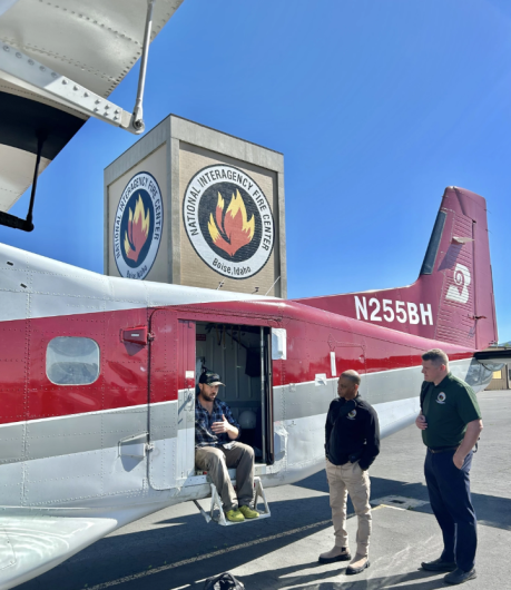 A person sits in the doorway of a small airplane talking to two people standing on the tarmac