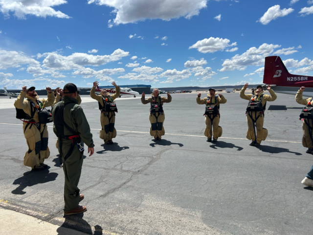 A group of people standing on airplane tarmac wearing tan jumpsuits and black gear strapped to their front look up with their hands raised.