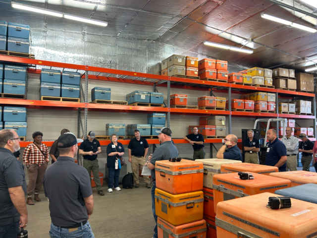 A group of people listen to a speaker in a warehouse with large cases on shelves