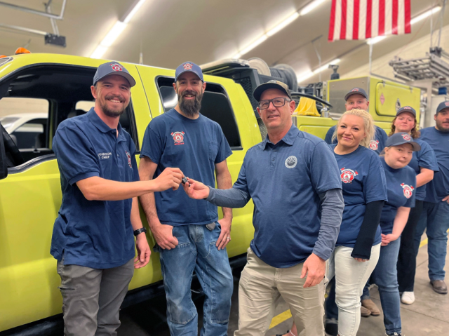 Caucasian men and women pose in front of a yellow truck, two people exchanging a set of keys